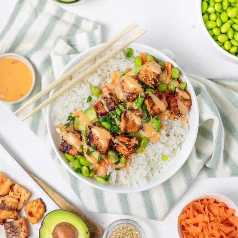 A rice bowl topped with seared salmon cubes, edamame, avocado and carrot, drizzled with bang bang sauce