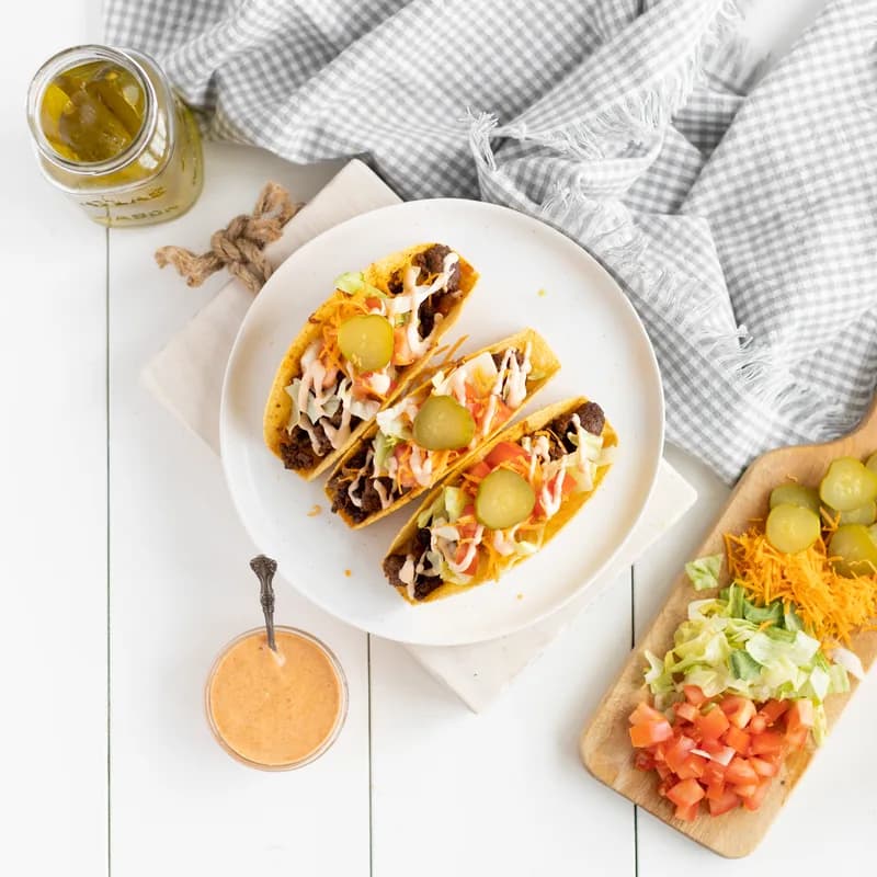 Three tacos filled with seasoned beef, shredded lettuce, cheese and pickles, with a bowl of special sauce beside them