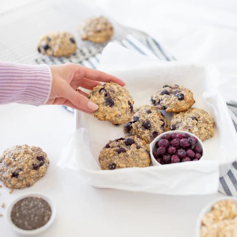 Oat breakfast cookies studded with blueberries on a lined tray, with a hand reaching in for one