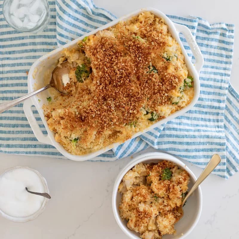 A broccoli and cheese casserole in a white baking dish under a golden breadcrumb crust, with a portion served alongside